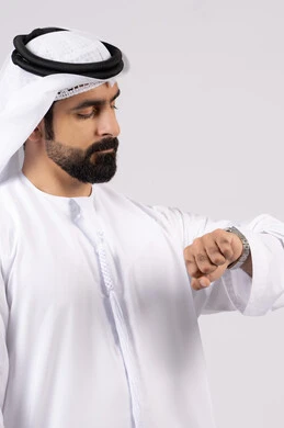 The concept of the importance of time and commitment to appointments, a portrait of an Emirati Gulf Arab man wearing a dakandura and ghutra looking at his personal watch with gestures of the importance of commitment to time and completing tasks and work accurately, white background