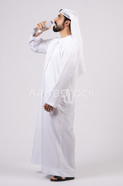 The importance of drinking pure water to maintain a fresh complexion, maintain a healthy immune system, eliminate toxins from the body, maintain and treat kidney health. Portrait of an Emirati Gulf Arab man wearing a kandura and ghutra drinking water from a plastic bottle, full body, white background.