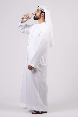 أهمية شرب الماء النقي للمحافظة على بشرة نضرة، المحافظة على سلامة الجهاز المناعي، التخلص من سموم الجسم، الحفاظ على صحة الكلى ومعالجتها، بورتريه لرجل عربي خليجي اماراتي  يرتدي الكندورة والغترة يقوم بشرب الماء م قارورة بلاستيكية، الجسم بالطول الكامل، خلفية بيضاء