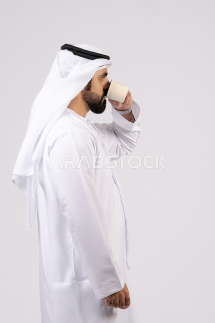 الاستمتاع بتناول المشروبات الساخنة والأعشاب الشتوية الدافئة،  تأثير الكافيين على نشاط الدماغ والذاكرة، بورتريه لرجل عربي خليجي اماراتي يرتدي الكندورة والغترة يحمل فنجان من القهوة،صورة الجسم بالطول الكامل، خلفية بيضاء