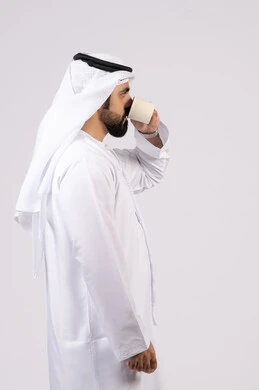 الاستمتاع بتناول المشروبات الساخنة والأعشاب الشتوية الدافئة،  تأثير الكافيين على نشاط الدماغ والذاكرة، بورتريه لرجل عربي خليجي اماراتي يرتدي الكندورة والغترة يحمل فنجان من القهوة،صورة الجسم بالطول الكامل، خلفية بيضاء