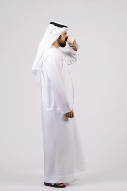 تأثير الكافيين على نشاط الدماغ والذاكرة، الاستمتاع بتناول المشروبات الساخنة والأعشاب الشتوية الدافئة، بورتريه لرجل عربي خليجي اماراتي يرتدي الكندورة والغترة يحمل فنجان من القهوة،صورة الجسم بالطول الكامل، خلفية بيضاء