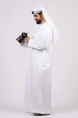 Professional photography using digital cameras, gestures and expressions of pleasure and happiness, portrait of a smiling young Emirati Arab man wearing a kandura and ghutra taking souvenir photos with a camera, practicing the hobby of photography, full body, white background