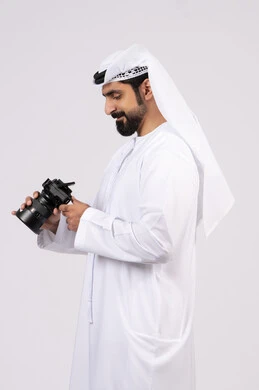 ايماءات وتعابير السرور والسعادة،بورتريه لشاب عربي خليجي اماراتي مبتسم يرتدي الكندورة والغترة يقوم بالتقاط صور تذكارية عبر كاميرا فوتوغرافية, ممارسة هواية التصوير, احتراف التصوير الفوتوغرافي باستخدام الكاميرات الرقمية, الجسم بالطول الكامل، خلفية بيضاء