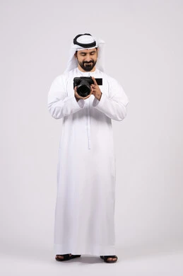 بورتريه لشاب عربي خليجي اماراتي مبتسم يرتدي الكندورة والغترة يقوم بالتقاط صور تذكارية عبر كاميرا فوتوغرافية, ممارسة هواية التصوير, احتراف التصوير الفوتوغرافي باستخدام الكاميرات الرقمية,ايماءات وتعابير السرور والسعادة، الجسم بالطول الكامل، خلفية بيضاء