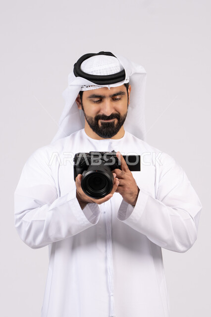 ممارسة هواية التصوير, احتراف التصوير الفوتوغرافي باستخدام الكاميرات الرقمية,ايماءات وتعابير السرور والسعادة،  بورتريه لشاب عربي خليجي اماراتي مبتسم يرتدي الكندورة والغترة يقوم بالتقاط صور تذكارية عبر كاميرا فوتوغرافية, الجسم بالطول الكامل، خلفية بيضاء