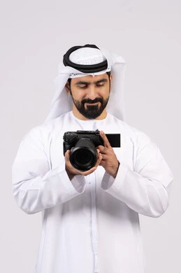 ممارسة هواية التصوير, احتراف التصوير الفوتوغرافي باستخدام الكاميرات الرقمية,ايماءات وتعابير السرور والسعادة،  بورتريه لشاب عربي خليجي اماراتي مبتسم يرتدي الكندورة والغترة يقوم بالتقاط صور تذكارية عبر كاميرا فوتوغرافية, الجسم بالطول الكامل، خلفية بيضاء