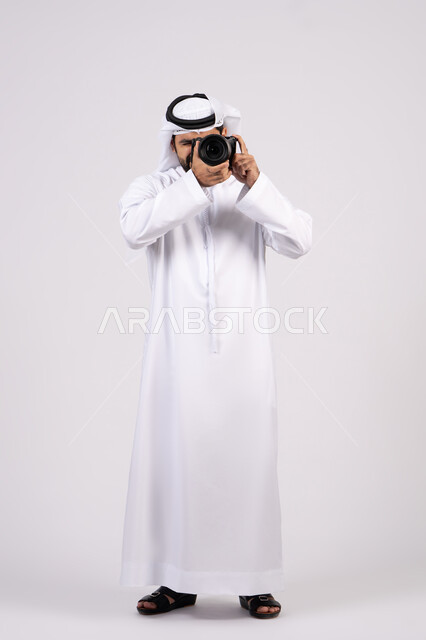 Professional photography using digital cameras, gestures and expressions of pleasure and happiness, practicing the hobby of photography, portrait of a smiling young Arab Emirati man wearing a kandura and ghutra taking souvenir photos with a camera, full body length, white background