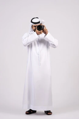 Professional photography using digital cameras, gestures and expressions of pleasure and happiness, practicing the hobby of photography, portrait of a smiling young Arab Emirati man wearing a kandura and ghutra taking souvenir photos with a camera, full body length, white background