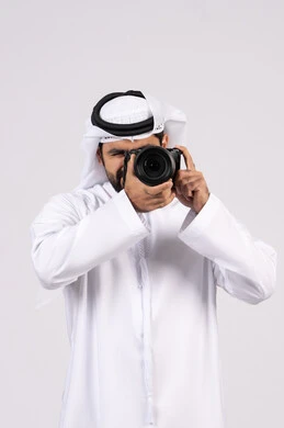 ايماءات وتعابير السرور والسعادة، احتراف التصوير الفوتوغرافي باستخدام الكاميرات الرقمية, ممارسة هواية التصوير, بورتريه لشاب عربي خليجي اماراتي مبتسم يرتدي الكندورة والغترة يقوم بالتقاط صور تذكارية عبر كاميرا فوتوغرافية, الجسم بالطول الكامل، خلفية بيضاء
