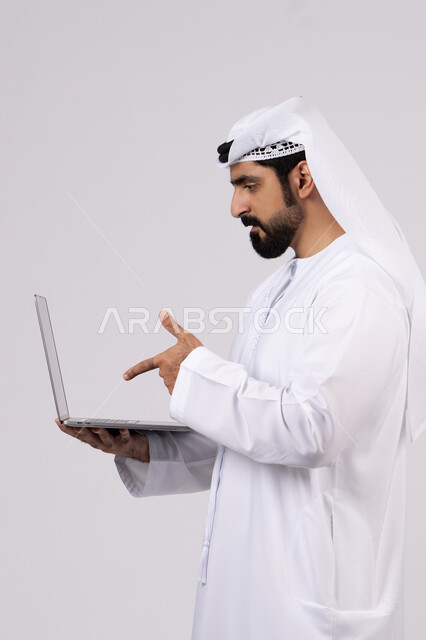 The concept of facilitating business and the ability to complete tasks remotely, managing and organizing business affairs through modern technical devices, integrating work with technology and technology, a portrait of a young Emirati Gulf Arab man wearing a kandura and ghutra, pointing to a laptop, full body, white background.