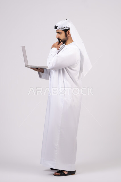 إدارة شؤون الأعمال وتنظيمها عبر الأجهزة التقنية الحديثة، دمج العمل مع التقنيات والتكنولوجيا ،مفهوم تسهيل الاعمال والقدرة على إنجاز المهام عن بعد، بورتريه لشاب عربي خليجي اماراتى يرتدي الكندورة والغترة يمسك بيده حاسوب محمول تظهر عليه علامات التفكير والحيرة، الجسم بالطول الكامل، خلفية بيضاء.