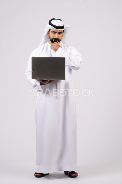 دمج العمل مع التقنيات والتكنولوجيا ، إدارة شؤون الأعمال وتنظيمها عبر الأجهزة التقنية الحديثة، مفهوم تسهيل الاعمال والقدرة على إنجاز المهام عن بعد، بورتريه لشاب عربي خليجي اماراتى يرتدي الكندورة والغترة يمسك بيده حاسوب محمول تظهر عليه علامات التفكير والحيرة، الجسم بالطول الكامل، خلفية بيضاء.