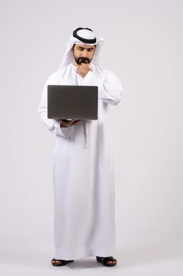 دمج العمل مع التقنيات والتكنولوجيا ، إدارة شؤون الأعمال وتنظيمها عبر الأجهزة التقنية الحديثة، مفهوم تسهيل الاعمال والقدرة على إنجاز المهام عن بعد، بورتريه لشاب عربي خليجي اماراتى يرتدي الكندورة والغترة يمسك بيده حاسوب محمول تظهر عليه علامات التفكير والحيرة، الجسم بالطول الكامل، خلفية بيضاء.