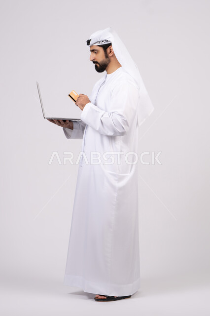 مفهوم الدفع والتسديد عن بعد عبر تطبيقات اللابتوب،استخدام البطاقة البنكية الالكترونية، تطور التكنولوجيا والأجهزة التقنية، بورتريه لرجل عربي اماراتي خليجي يرتدي الكندورة والغترة يعمل على جهاز الحاسوب المحمول، خلفية بيضاء