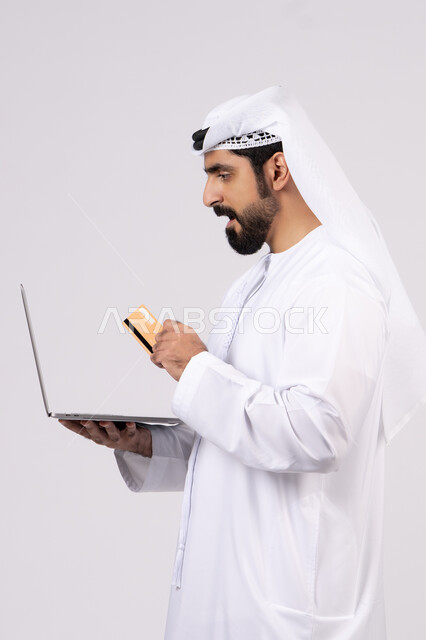 استخدام البطاقة البنكية الالكترونية، مفهوم الدفع والتسديد عن بعد عبر تطبيقات اللابتوب، تطور التكنولوجيا والأجهزة التقنية، بورتريه لرجل عربي اماراتي خليجي يرتدي الكندورة والغترة يعمل على جهاز الحاسوب المحمول، خلفية بيضاء