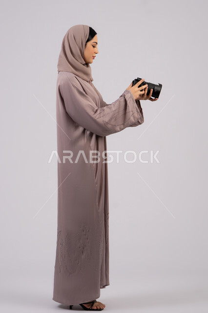 Professional photography, using high-resolution digital cameras, using modern technical devices and technology, portrait of an Emirati Gulf Arab woman wearing a hijab and abaya taking a souvenir photo with a camera, a Saudi photographer practicing the hobby of photography with expressions of pleasure, a female job and profession, white background