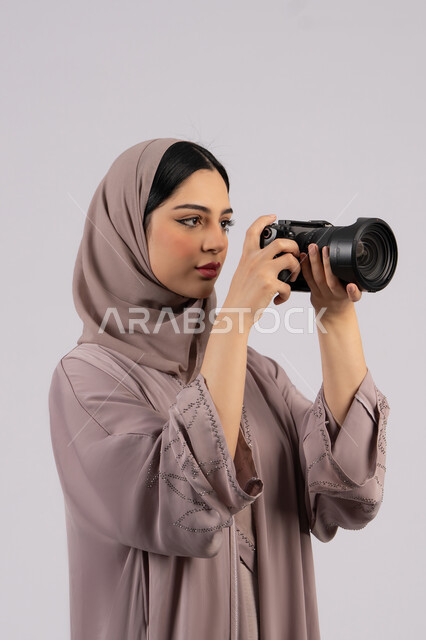 Professional photography using high-resolution digital cameras, women's job and profession, portrait of an Emirati Gulf Arab woman wearing a hijab and abaya taking a souvenir photo with a camera, a Saudi photographer practicing the hobby of photography with expressions of pleasure, white background