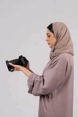 بورتريه من الجانب لإمراة عربية خليجية اماراتية ترتدي الحجاب والعباءة تقوم بالتقاط صورة تذكارية عبر كاميرا فوتوغرافية,  مصورة سعودية تمارس هواية التصوير بتعابير السرور،وظيفة ومهنة نسائية, احتراف فن التصوير الفوتوغرافي باستخدام كاميرات رقمية عالية الدقة, خلفية بيضاء
