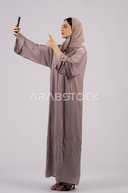 Quality and excellence gestures, technology and modern technology, having fun on mobile with friends, browsing social networking sites, portrait of a veiled Emirati Gulf Arab woman wearing an abaya using a mobile phone, Emirati woman taking a selfie, full body, white background