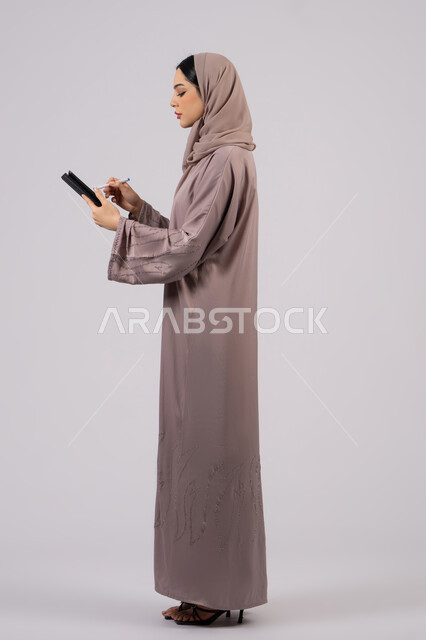 النظر بتعابير الاندماج , دمج التقنيات الحديثة والتكنولوجيا في الحياة العملية،بورتريه لإمرأة عربية خليجية سعودية محجبة ترتدي العباءة تقوم باستخدام قلم الجهاز اللوحي، سيدة اماراتية تقوم بتسجيل وتدوين البيانات والملاحظات، صورة للجسم بالكامل، خلفية بيضاء