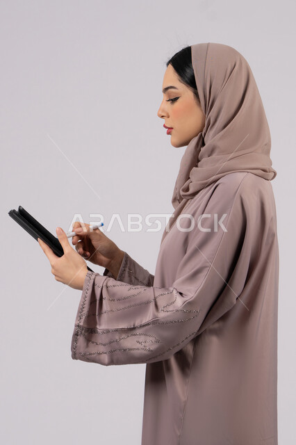 دمج التقنيات الحديثة والتكنولوجيا في الحياة العملية، النظر بتعابير الاندماج , بورتريه لإمرأة عربية خليجية سعودية محجبة ترتدي العباءة تقوم باستخدام قلم الجهاز اللوحي ،سيدة اماراتية تقوم بتسجيل وتدوين البيانات والملاحظات، صورة للجسم بالكامل، خلفية بيضاء