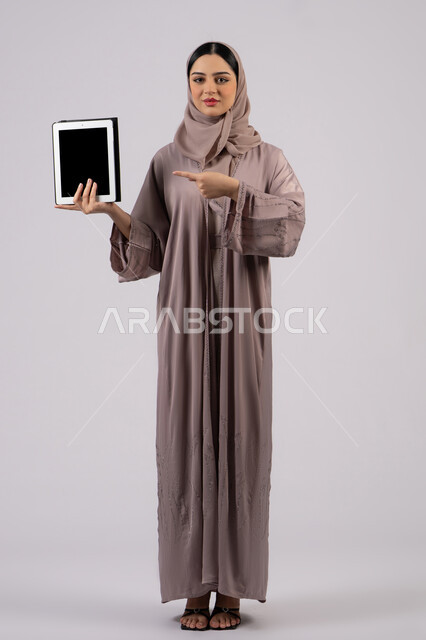Gestures of interaction and integration, integration of modern technologies in practical life, portrait of a veiled Saudi Arabian Gulf woman wearing an abaya using a tablet, an Emirati woman displaying a blank black screen, a full body image, a white background