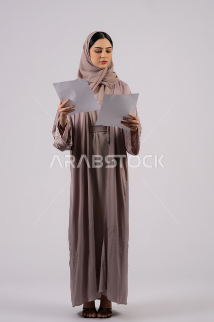 تدوين المهام اليومية ،شابه سعوديه تقوم بالبحث العلمي، قراءة الملاحظات والمعلومات ، متابعة الخطط والمشاريع ،أوراق خاصة بالعمل,بورتريه لامراة عربيه خليجيه اماراتيه محجبه ترتدي العباءة السوداء تحمل بيديها ورقة بيضاء ،  خلفية بيضاء