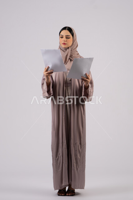 Following up on plans and projects, work papers, writing down daily tasks, portrait of a veiled Arab Gulf Emirati woman wearing a black abaya holding a white paper and a pen, a young Saudi woman doing scientific research, reading notes and information, white background