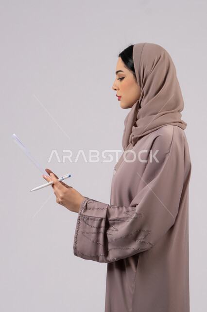 تدوين المهام اليومية ، أوراق خاصة بالعمل, بورتريه لامراة عربيه خليجيه اماراتيه محجبه ترتدي العباءة السوداء تحمل بيديها ورقة بيضاء وقلم، شابه سعوديه تقوم بالبحث العلمي، قراءة الملاحظات والمعلومات ، متابعة الخطط والمشاريع ، خلفية بيضاء