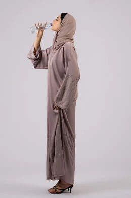 Side portrait of a veiled Emirati Gulf Arab woman wearing an abaya drinking from a small plastic bottle of water, a Saudi woman feeling thirsty and dehydrated, maintaining the health of the body's immune system, regularly drinking water, white background