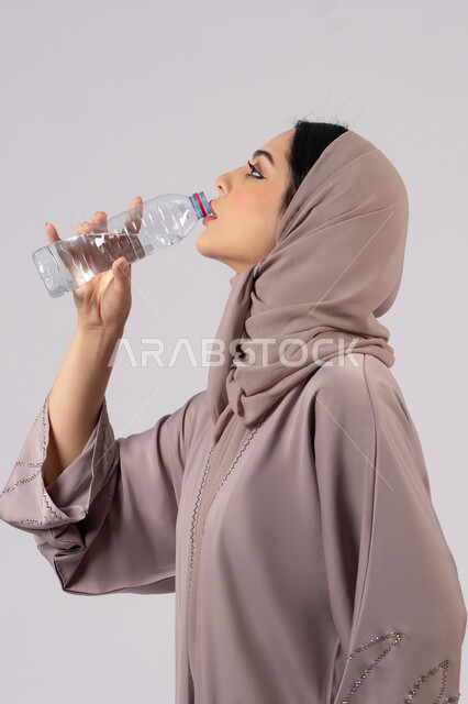 المحافظة على سلامة الجهاز المناعي للجسم , المداومة على شرب المياه ،بورترية من الجانب لامراة عربية خليجية امارتية محجبة ترتدي العباءة تقوم بالشرب من زجاجة ماء بلاستيكية صغيرة, سيدة سعودية تشعر بالعطش والجفاف , خلفية بيضاء