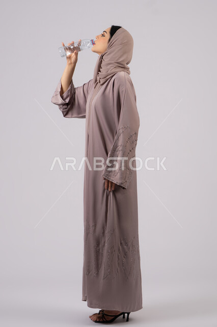 Drinking water regularly maintains the health of the body's immune system. Portrait of a veiled Emirati Gulf Arab woman wearing an abaya drinking from a small plastic water bottle. A Saudi woman feels thirsty and dehydrated. White background.
