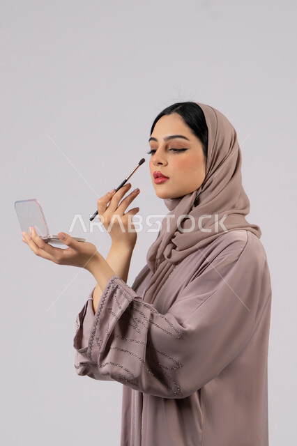 مفهوم الانوثه والاهتمام بالمظهر الخارجي، سيدة اماراتية تستخدم أدوات تجميل نسائيه عصريه، مكياج وميكاب تتوريال، صالون نسائي راقي، التسويق لمنتجات التجميل ،بورتريه لفتاه عربيه سعودية خليجيه ترتدي العباءة والحجاب تحمل بيدها فرشاة توريد الخدود