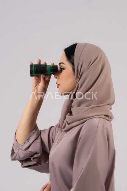 حب الاستكشاف والمغامرة، ايماءات الدهشه و الاستغراب, التخطيط المستقبلي للأهداف و الطموحات، شابة سعودية تقوم برؤية الاشياء البعيدة عبر التلسكوب، بورتريه من الجانب لامرأة عربية خليجية اماراتية محجبة ترتدي العباءة والحجاب تنظر من خلال منظار