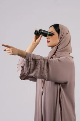 Pointing to something, future planning for goals and ambitions, a young Saudi woman seeing distant things through a telescope, a side portrait of a veiled Emirati Gulf Arab woman wearing an abaya and hijab looking through binoculars with gestures of surprise and astonishment, love of exploration and adventure