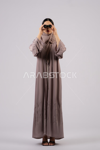 Trying to see something, portrait of a veiled Emirati Gulf Arab woman wearing an abaya and hijab looking through a telescope with gestures of astonishment and surprise, love of exploration and adventure, future planning for goals and ambitions, a young Saudi woman seeing distant things through a telescope