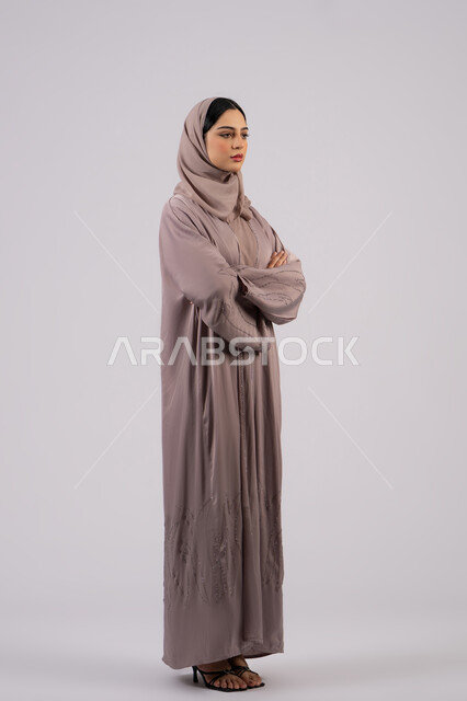 The concept of elegance and interest in outward appearance, a Saudi girl standing with crossed hands and self-confidence, a side portrait of an Arab Gulf Emirati woman wearing an abaya and hijab looking at something, full body length, white background