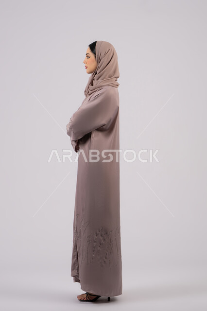Expressions of seriousness and concentration, side portrait of an Emirati Gulf Arab woman wearing an abaya and hijab looking at something, elegance and interest in outward appearance, the concept of femininity and softness, a Saudi girl standing with crossed hands and self-confidence, full body length, white background