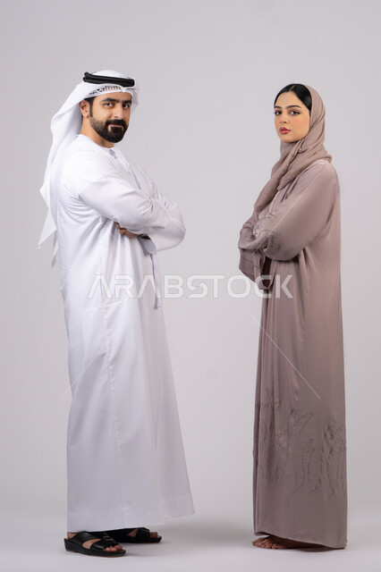 مفهوم الأناقة والاهتمام بالمظهر الخارجي، الوقوف بايدي متقاطعة وثقة بالنفس،  تعابير الجدية والتركيز، بورتريه  لزوجان عربيان خليجيان إماراتيان يرتديان الزي الاماراتي ينظران إلى شئ ما، الطول الكامل للجسم، خلفية بيضاء