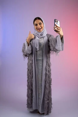 إيماءات الإعجاب والرضا، الشعور بالقبول والنظر إلى الكاميرا،  سيده سعودية تقوم بألتقاط صور سيلفي, اجراء مكالمة فيديو من خلال الهاتف الجوال، التواصل والتحدث مع الأهل والأصدقاء, بورتريه لإمرأة عربية خليجية اماراتية ترتدي العباءة الملونه تمسك بيدها الهاتف المحمول، الجسم بالطول الكامل ، خلفية ملونه