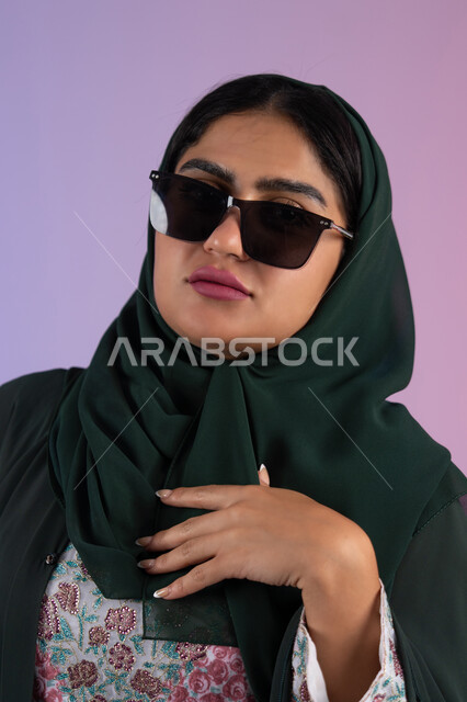 The concept of taking care of one's appearance, interest in fashion and elegance, a close-up portrait of an Emirati Gulf Arab woman wearing a colorful abaya and sunglasses, looking with confidence and self-esteem, a young Saudi woman standing straight and looking at the camera with gestures of coquettishness and femininity, a colorful background