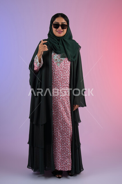 Interest in fashion and elegance, the concept of taking care of one's appearance, a portrait of an Emirati Gulf Arab woman wearing a colorful abaya and sunglasses, looking with confidence and self-esteem, a young Saudi woman standing straight and looking at the camera with gestures of coquettishness and femininity, full-length body, colorful background