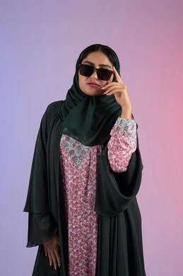 Modern and elegant veiled fashion, the concept of taking care of one's appearance, interest in fashion and elegance, a close-up portrait of an Emirati Gulf Arab woman wearing a colorful abaya and sunglasses, looking with confidence and self-esteem, a young Saudi woman standing straight and looking at the camera with gestures of coquettishness and femininity, a colorful background