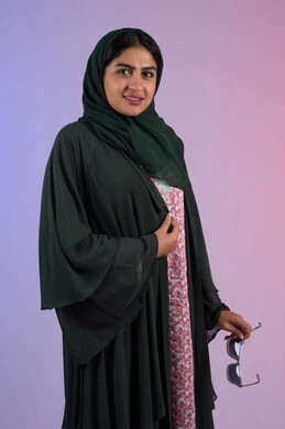 السرور والثقة بالنفس، مفهوم العناية بالجمال والإطلالة المتميزة، بورترية مقرب لسيدة عربية خليجية اماراتية مبتسمه محجبة ترتدي العباءه الملونه تمسك بيدها النظارة الشمسية ، سيدة سعودية تنظر الى الكاميرا بايماءات السعادة و السرور، خلفية ملونه