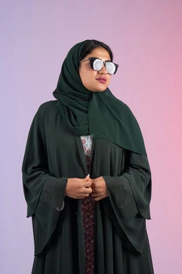 Self-care and outward appearance, the concept of femininity and elegance, beauty care and a distinguished appearance, a side portrait of a smiling veiled Emirati Gulf Arab woman wearing a colorful abaya and sunglasses, a Saudi woman standing with gestures of femininity and confidence, colorful background