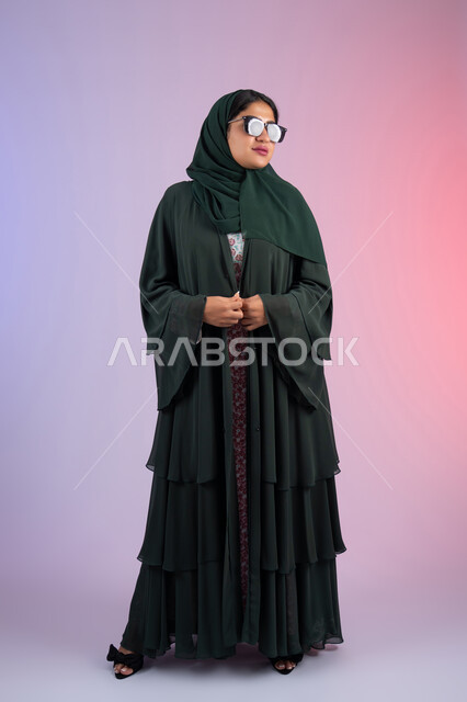 مفهوم الانوثه و الشياكة ، العناية بالجمال والإطلالة المتميزة، الأناقة والاهتمام بالمظهر الخارجي، بورترية من الجانب لسيدة عربية خليجية اماراتية مبتسمه محجبة ترتدي العباءه الملونه و النظارة الشمسية ، امراة سعودية تقف بايماءات الانوثه و الثقه، الجسم بالطول الكامل، خلفية ملونه