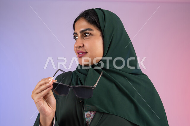 مفهوم الأنوثة والنعومة، ملابس محجبات عصرية راقية، بورتريه مقرب لامرأة عربية خليجية اماراتية ترتدي الحجاب والعباءة تمسك بيديها النظارة الشمسية تقف باستقامة بتعابير الثقة بالنفس، سيدة سعودية تنظر بإيماءات البشاشة والإعجاب، خلفية ملونه