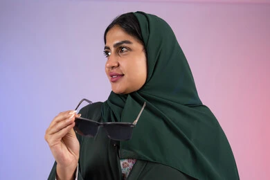 مفهوم الأنوثة والنعومة، ملابس محجبات عصرية راقية، بورتريه مقرب لامرأة عربية خليجية اماراتية ترتدي الحجاب والعباءة تمسك بيديها النظارة الشمسية تقف باستقامة بتعابير الثقة بالنفس، سيدة سعودية تنظر بإيماءات البشاشة والإعجاب، خلفية ملونه