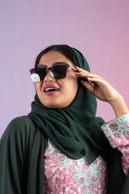 Femininity and beauty gestures, Emirati woman wearing elegant abaya and contemporary black glasses, close-up portrait of veiled Saudi Arabian Gulf woman trying on new sunglasses, interest in outward appearance and elegance, colorful background